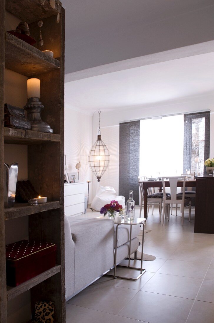 Rustic wooden shelving and view into bright, open-plan interior with modern couch and dining table with white, vintage-effect chairs