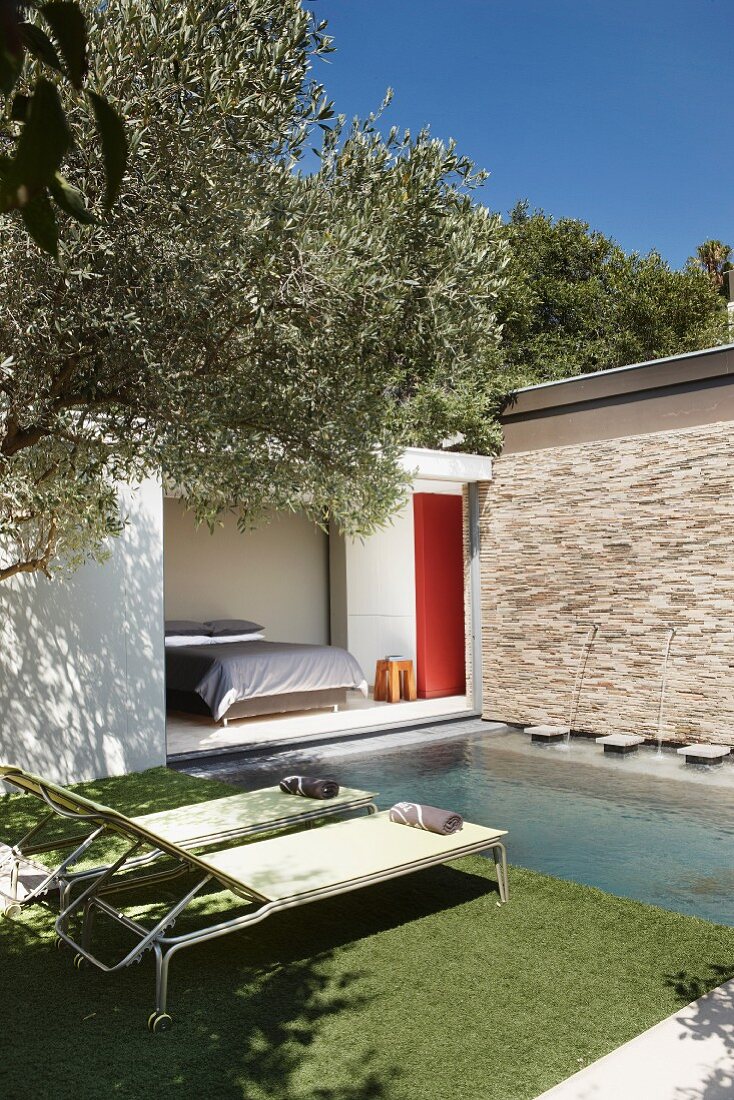 Pool in front of a bedroom with an open sliding door; in the foreground, two sun loungers on artificial turf