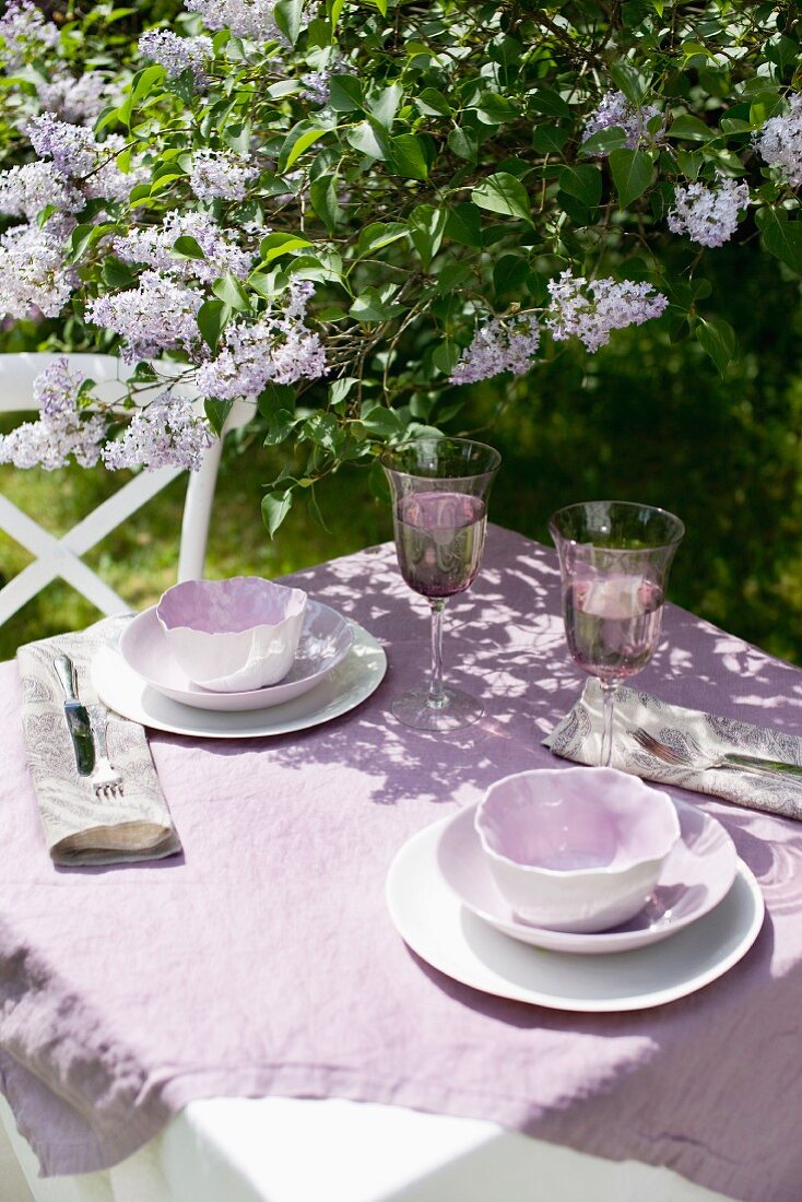 Sonnenbeschienener Gartentisch unter blühendem Flieder mit violetter Leinentischdecke und Porzellangeschirr im selben Farbton