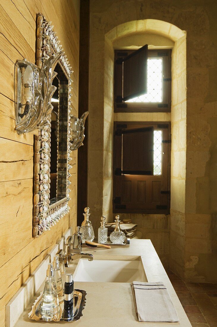 Ornate mirror flanked by sconce lamps on wooden wall above perfume bottles on washbasin; window niche with half-open interior shutters in background