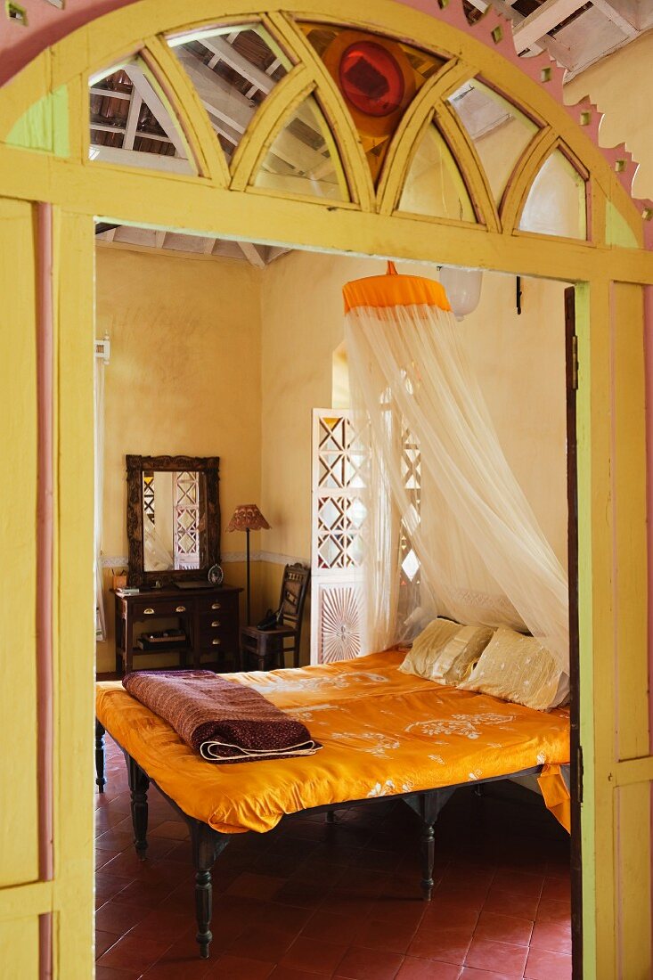 View of double bed with yellow bed linen through open door with carved archway of yellow-painted wood