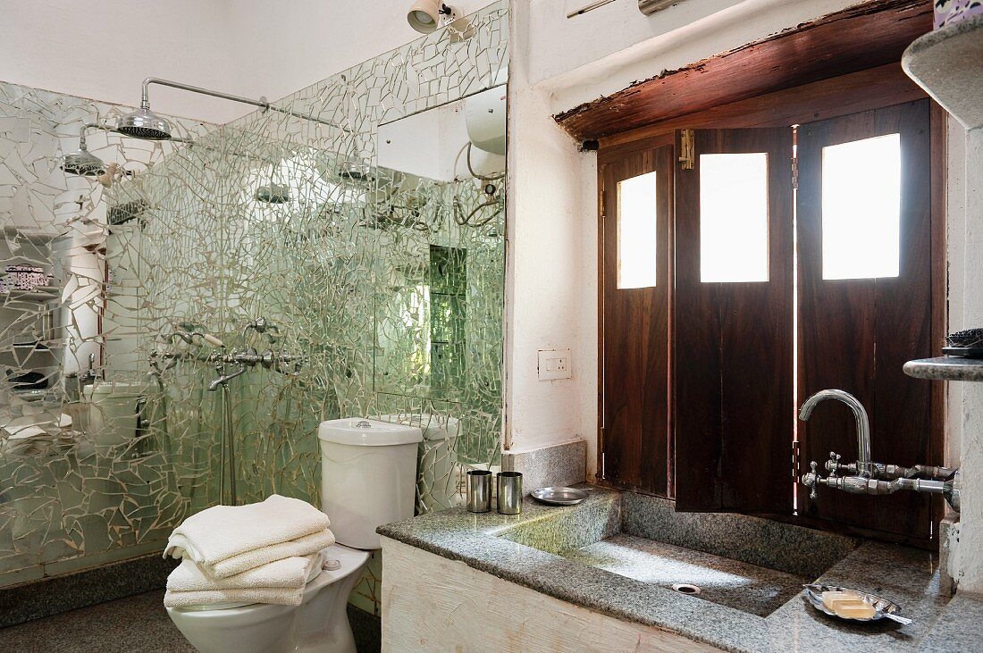 Washstand below window with interior wooden shutters and shower area with mirrored mosaic wall panels