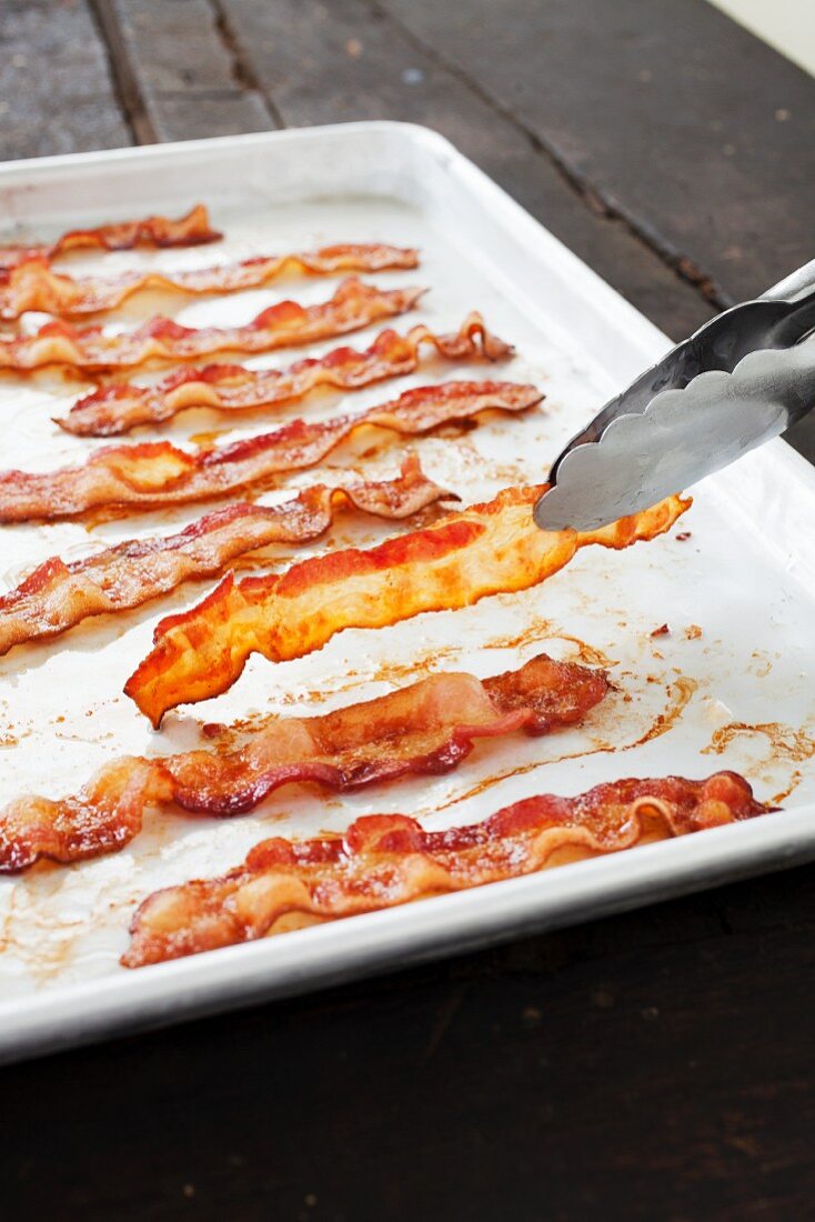 Tongs Flipping a Strip of Bacon on a Baking Sheet