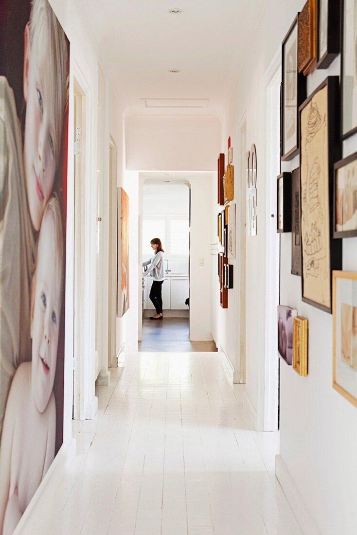 White wooden floor in narrow hallway with large painting and framed pictures on walls; woman in kitchen in background