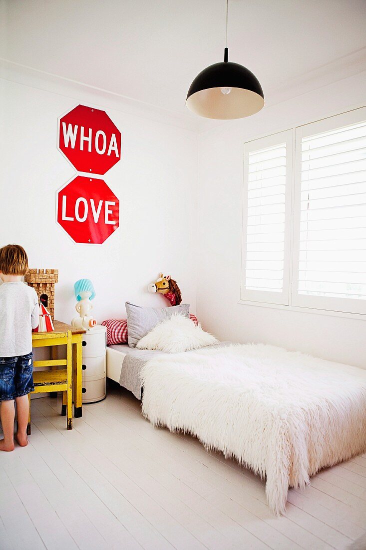 Child's bedroom in white - bed with flokati bedspread below window and little boy standing in front of knight's castle on table