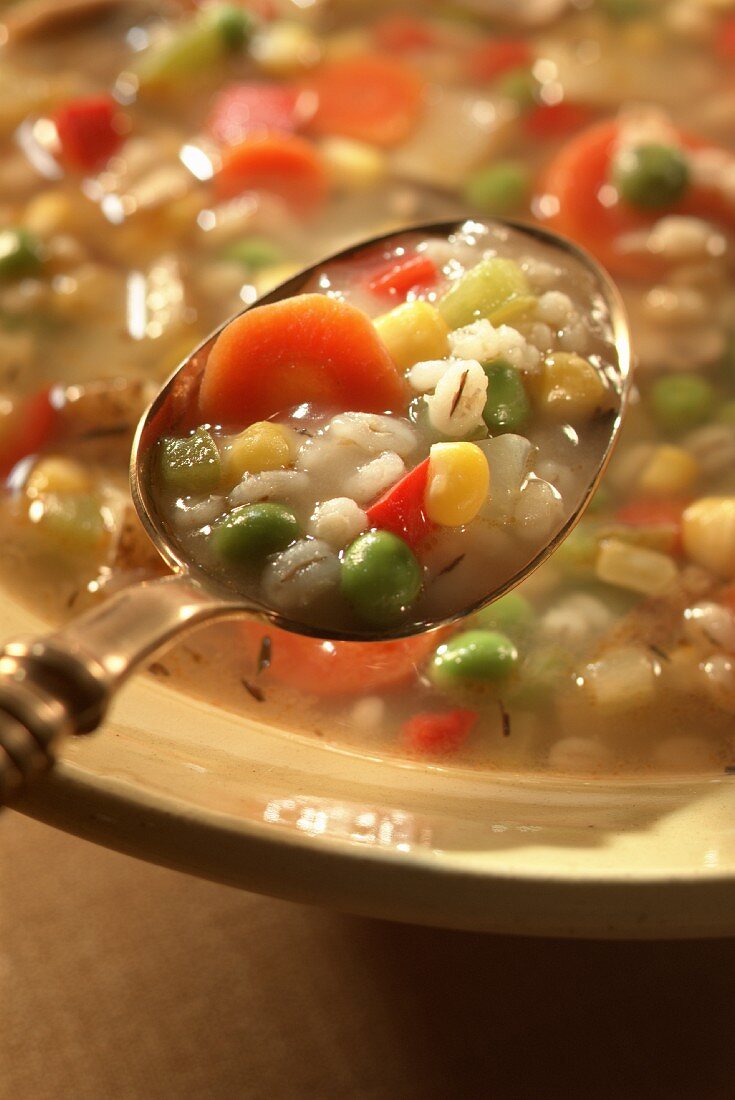 Graupensuppe mit Erbsen, Karotten und Mais auf einem Löffel und in einem Suppenteller
