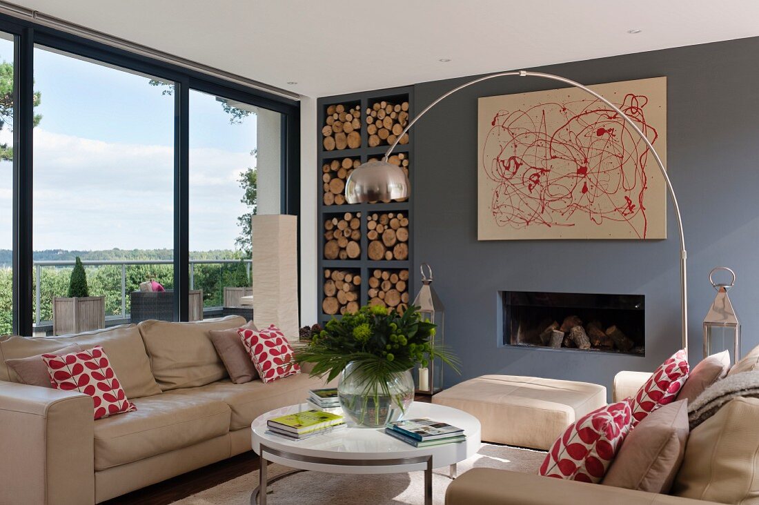 Beige sofa set and designer lamp above coffee table in front of fireplace in wall painted blue-grey; shelves of firewood