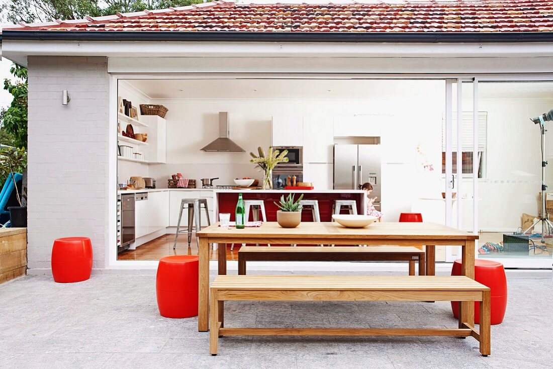 Möblierter Terrassenbereich mit roten fassförmigen Sitzhockern vor geöffneter Fensterfront; vollständiger Blick in den überwiegend weißen Küchenwohnbereich des Bungalows