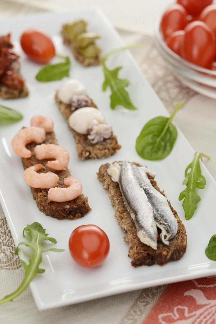 Slices of bread with sardines, shrimps and squid on a rectangular platter with lettuce leaves and cherry tomatoes