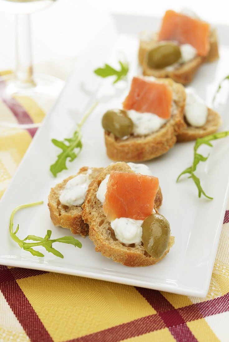 Canapés mit Kräuterfrischkäse, Räucherlachs und grüner Olive, auf rechteckiger weißer Platte