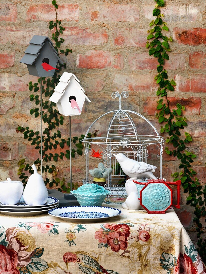 Stylised fruit on plate and china bird in front of birdcage on table against brick wall