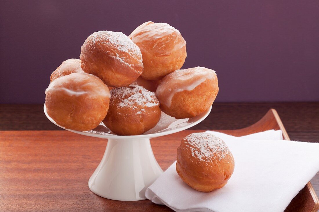 Several doughnuts coated with sugar glaze or icing sugar, on a cake stand against a purple background