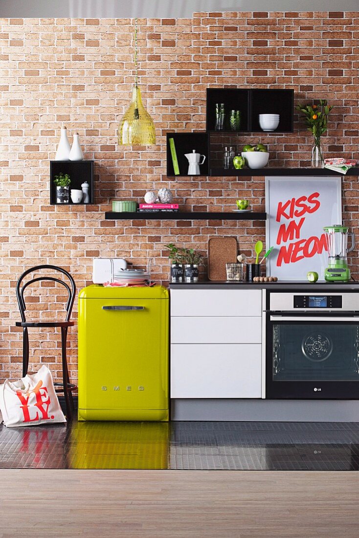 Electric kitchen appliances and white kitchen base unit in a row below various items of crockery on black shelves on brick wall