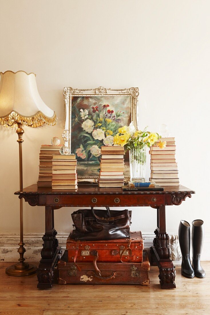 Stacked books and oil painting leaning on wall on top of antique desk above leather suitcases next to Biedermeier standard lamp