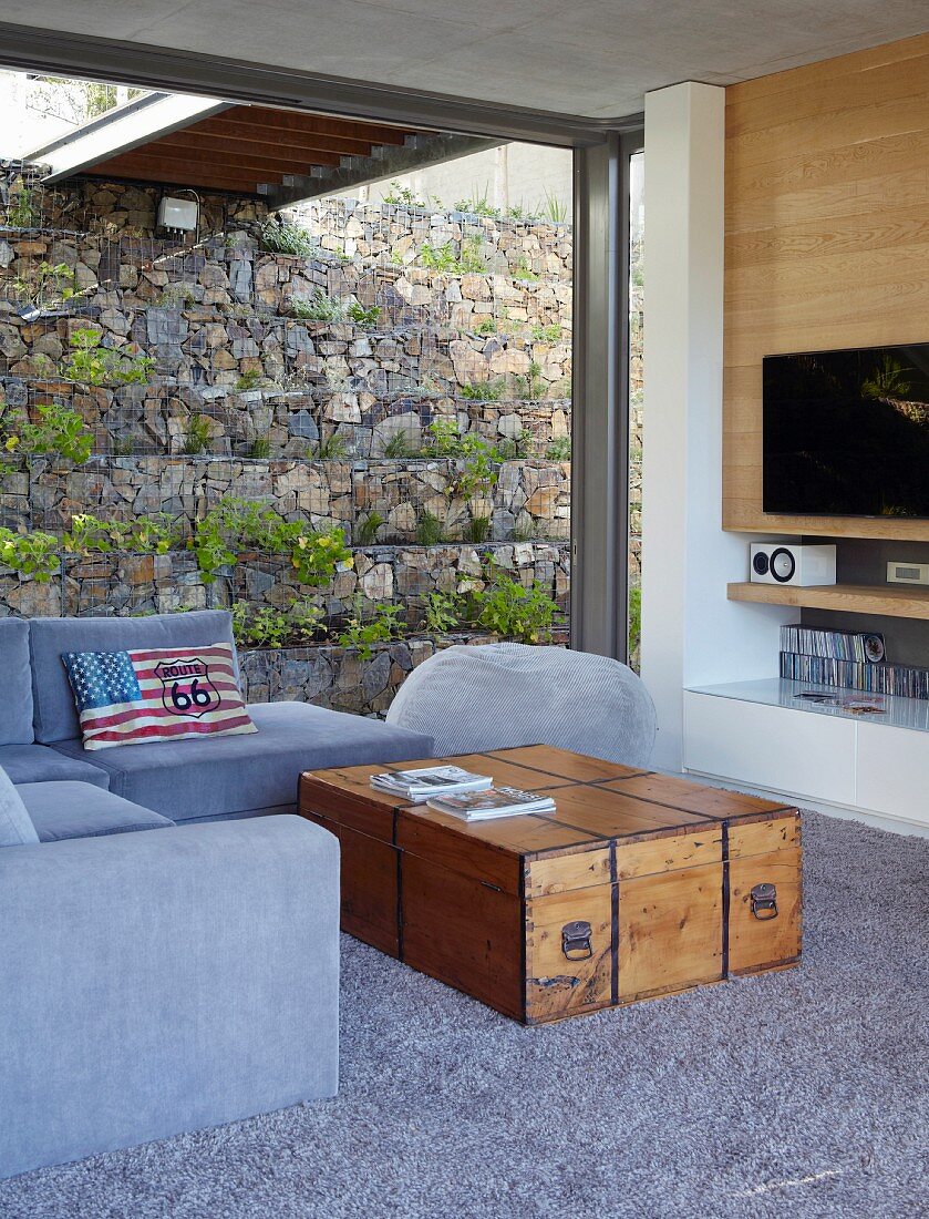 Daylight interior in house built into slope with planted gabion wall; vintage wooden trunk used as coffee table for seating area in shades of grey