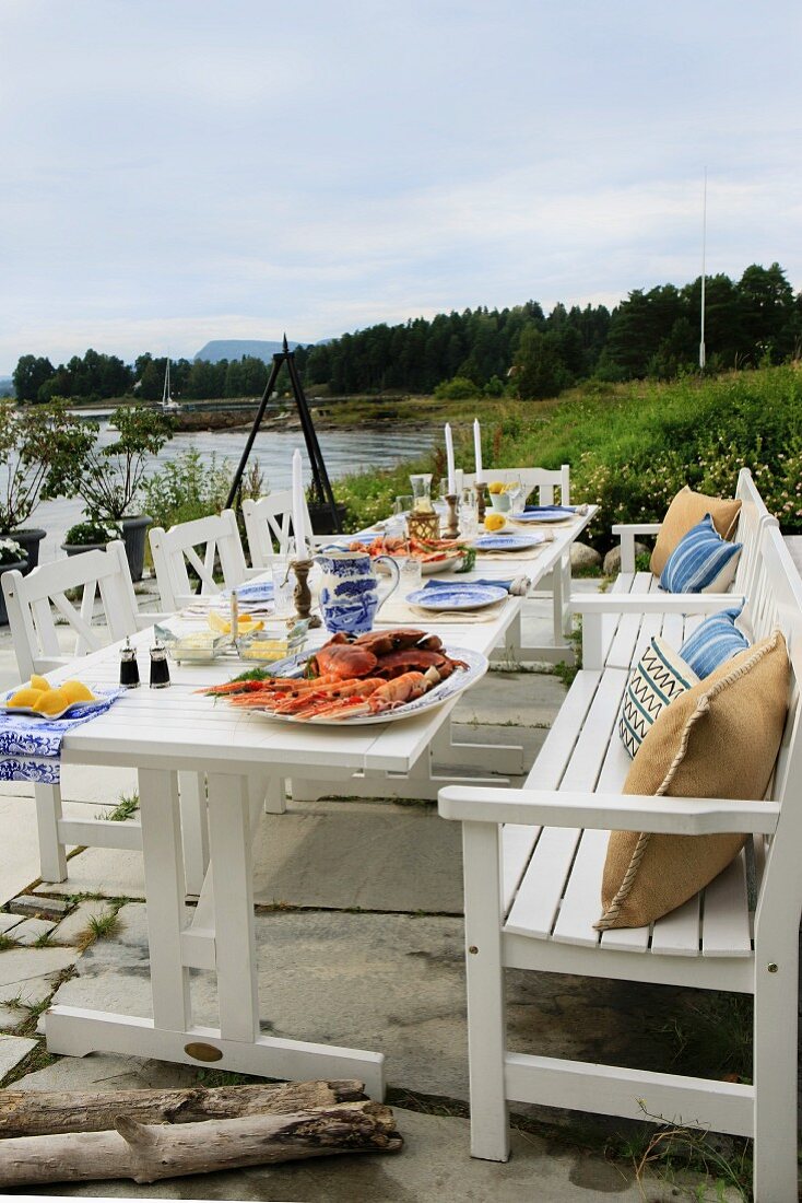 Festlich gedeckte Gartentafel mit robusten, weissen Holzmöbeln im Landhausstil auf Terrasse am Wasser