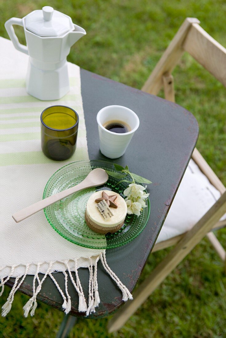 An espresso pot, mug of coffee and a glass plate with a sweet pastry on a garden table