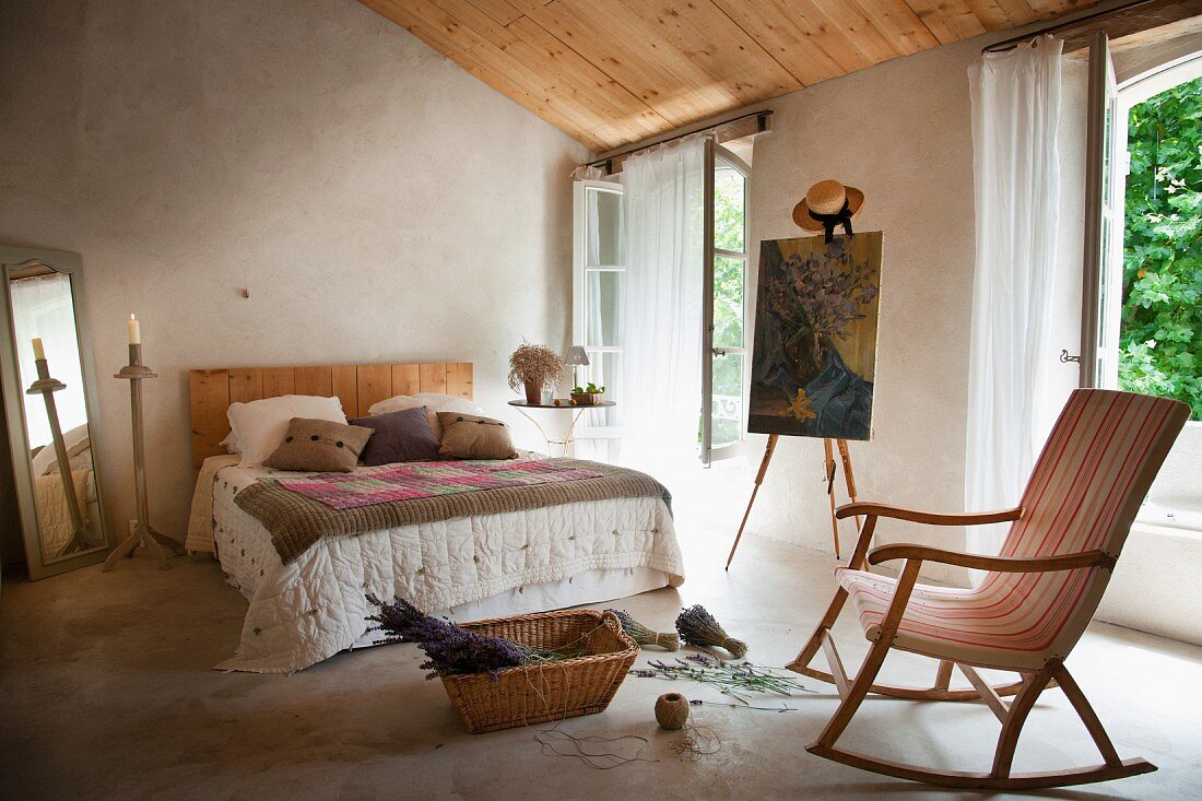 Korb mit Lavendelblüten vor einem Doppelbett, Staffelei mit Gemälde und ein Schaukelstuhl