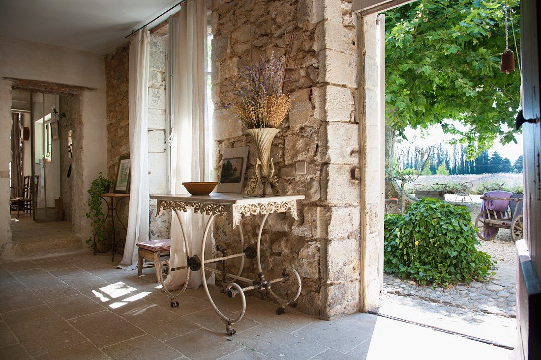 Zierlicher, alter Metalltisch vor rustikaler Natursteinwand und Blick durch eine offene Tür auf den Garten