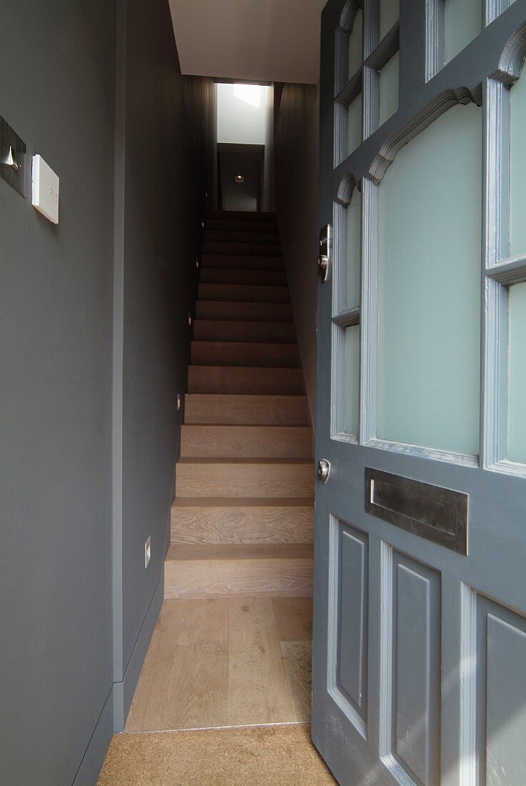 View through open front door into grey-painted stairwell with narrow wooden staircase