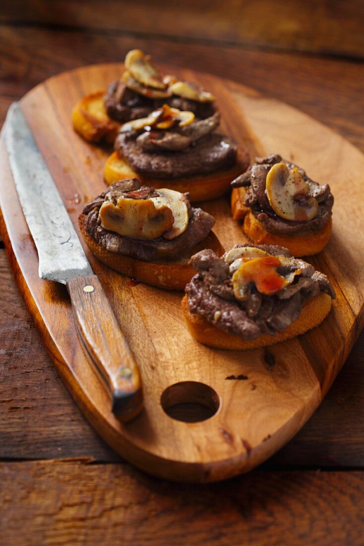 Toasted baguette slices topped with tournedos rossini on a wooden board with a knife