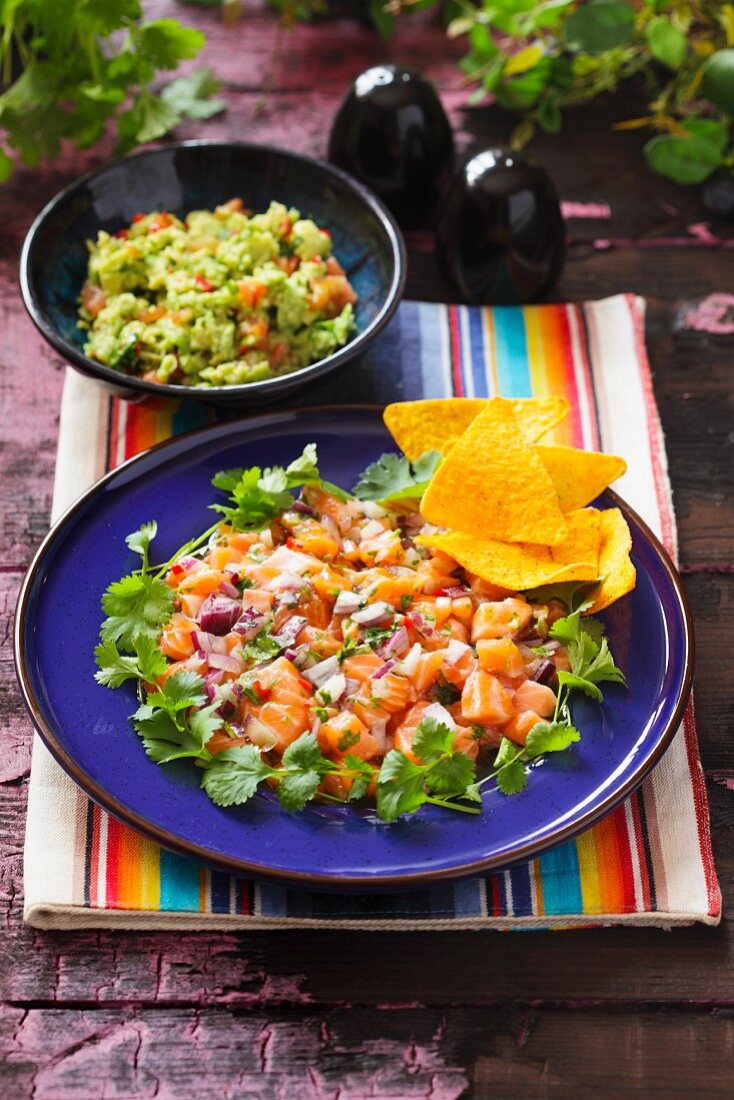 Ceviche as a main course on a blue plate with coriander and tortilla chips and a bowl of guacamole in the background