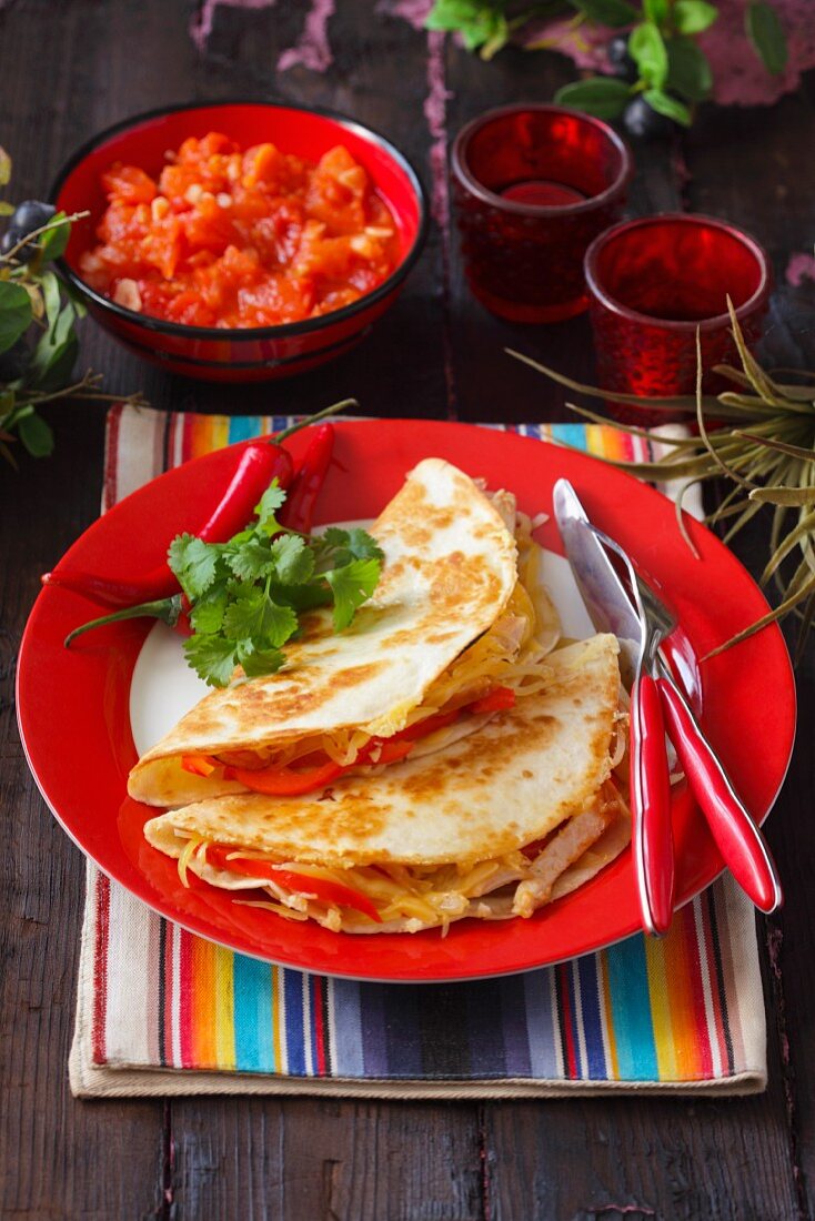 Two tortillas with chicken strips on a red plate on a colourfully striped placemat with a chilli dip in the background