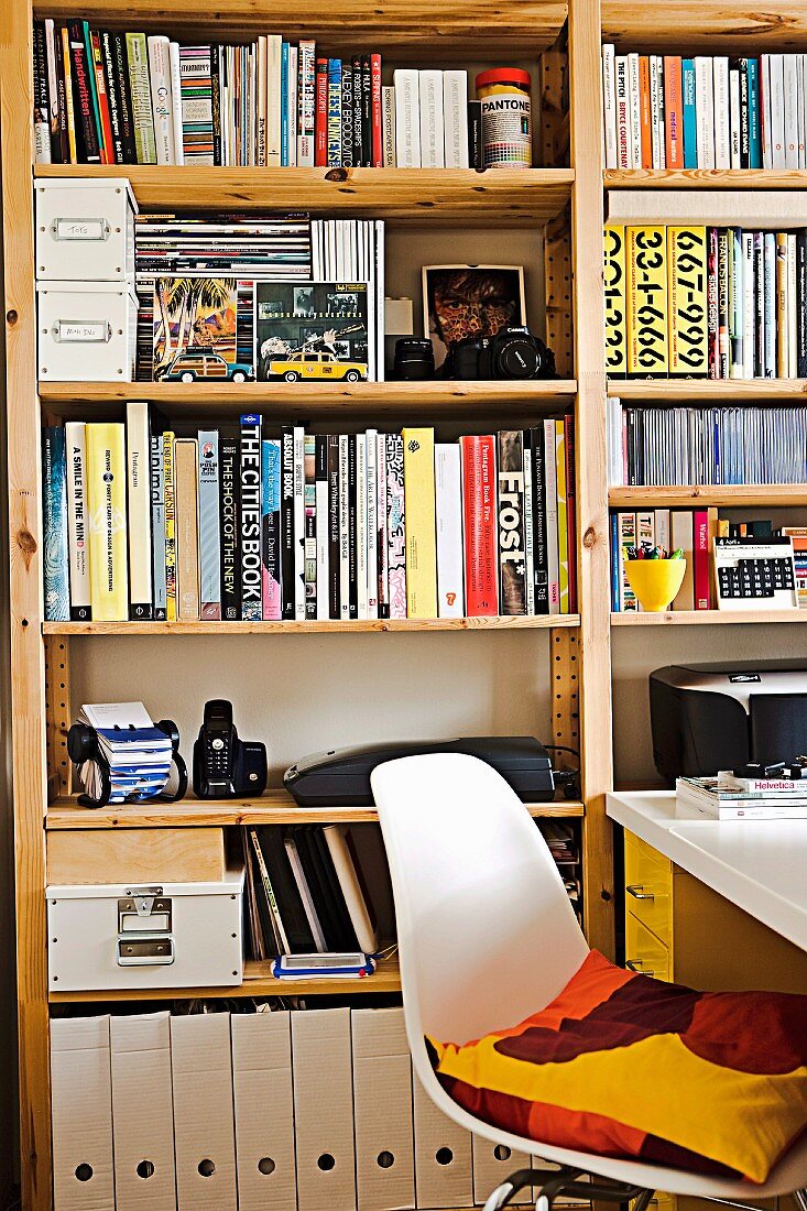 Full shelf wall in study with white shell chair and white desk; a colorful pillow on the chair