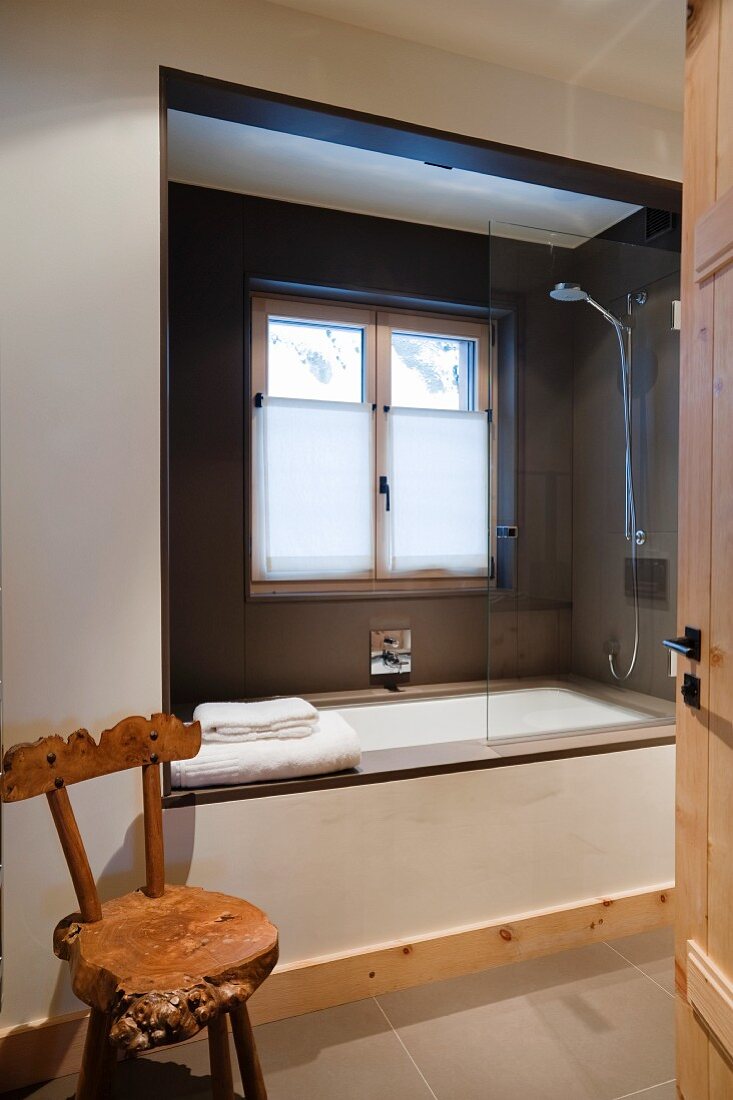 Blick durch offene Tür auf rustikalen Holzstuhl und eingebauter moderner Badewanne in Fensternische