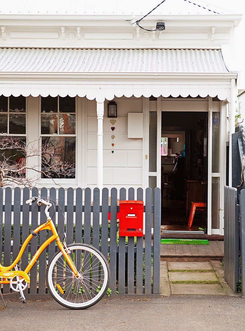 Entrance of white-painted cottage with porch; yellow bicycle leaning on grey garden fence with red letter box