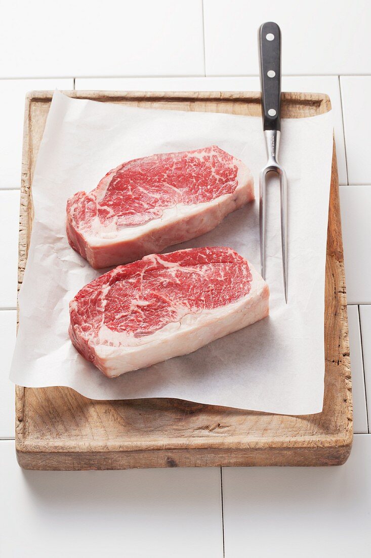 Two entrecote on piece of parchment paper on a wooden board with a carving fork