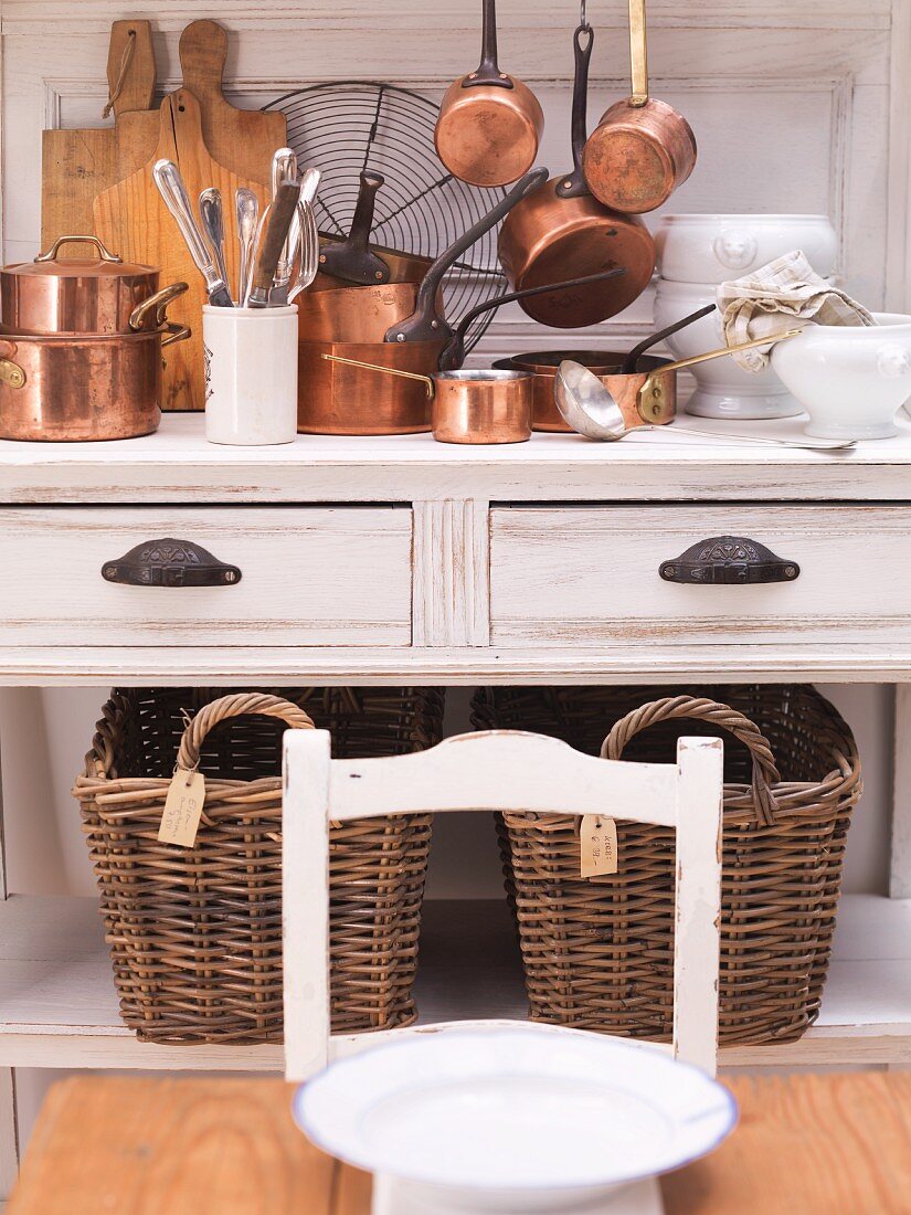 Shiny copper pots on a country house-style kitchen buffet with two large wicker baskets underneath