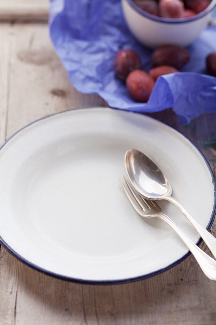 A fork and a spoon on a white plate with damsons in the background