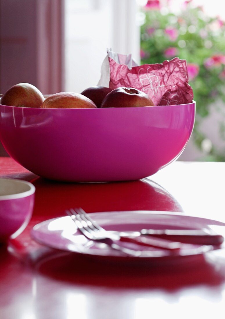 Cheerful mixture of colours - pink plastic bowl of apples and blurred, matching place setting in foreground on red table