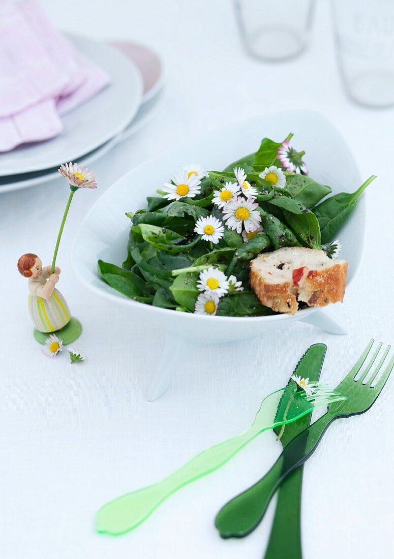 Grünes Plastik Besteck und Mädchenfigur neben Schale mit Spinatsalat und Gänseblümchen