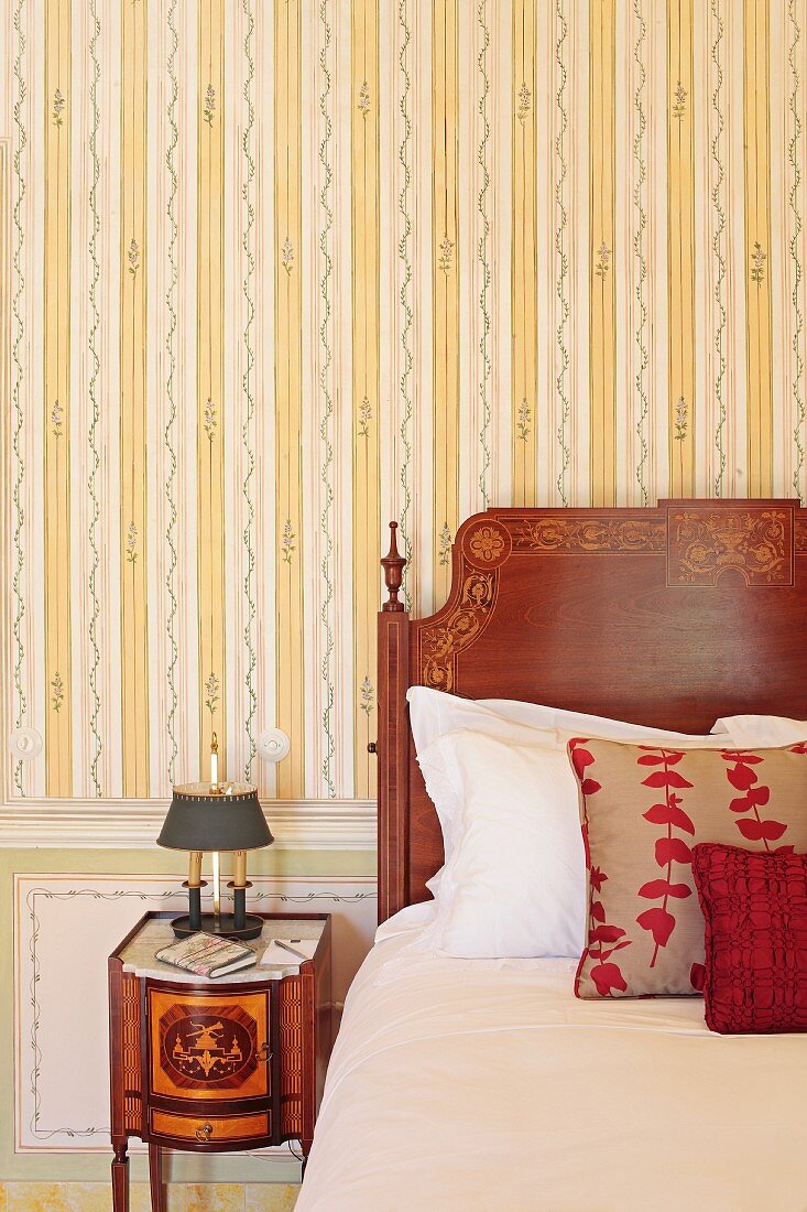 Detail of wooden bed headboard with marquetry veneer next to antique bedside table against striped wallpaper