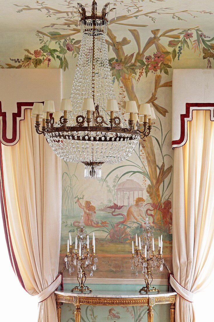 Chandelier with small white lampshades in front of painted wall and candlesticks on gilt console table