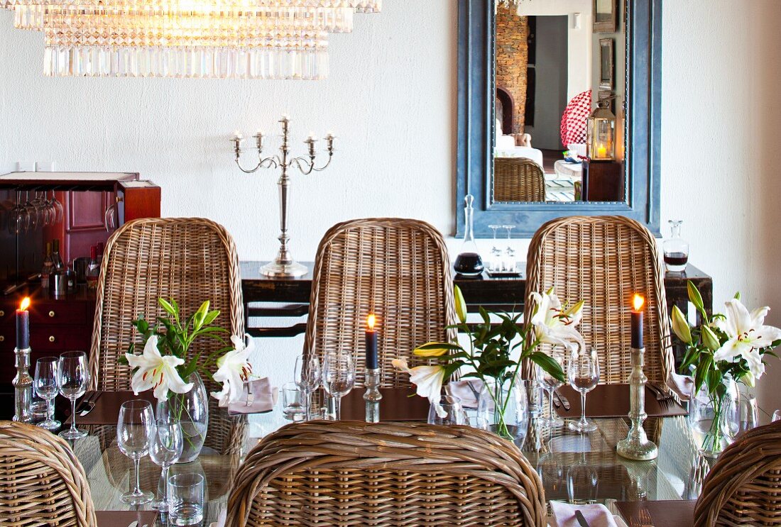 Candlesticks and arrangements of lilies on lustrous glass table surrounded by wicker chairs with curved backs