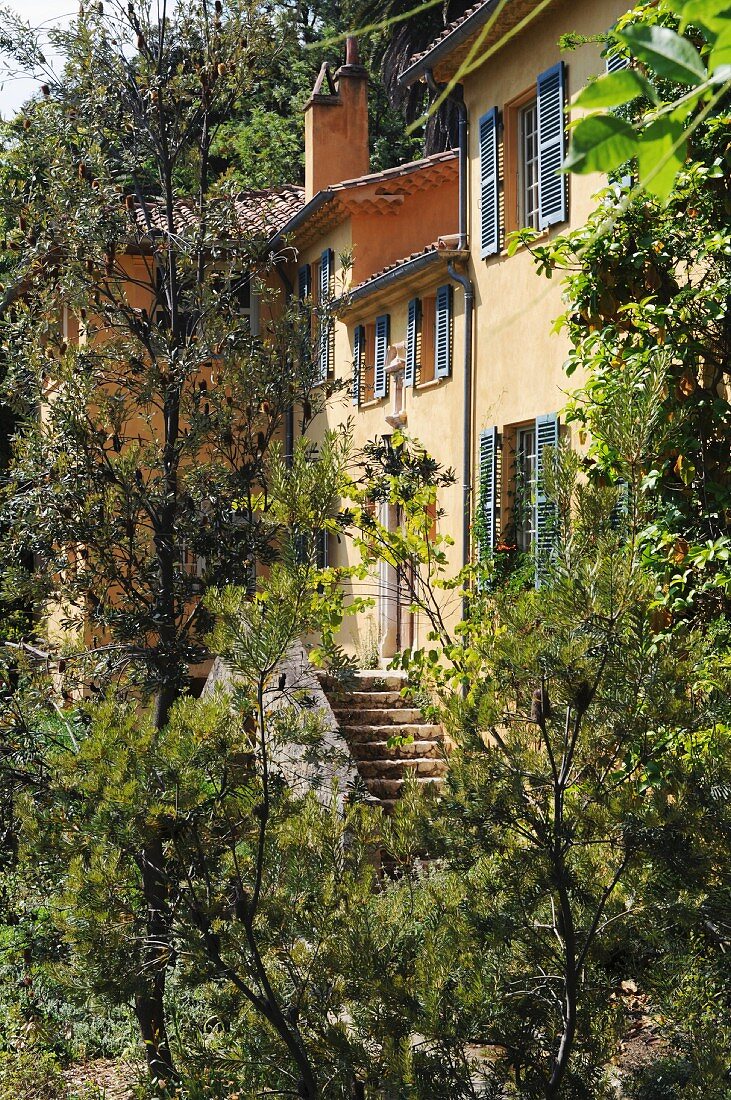 Yellow country house with grey shutters and terracotta tiled roof hidden behind trees