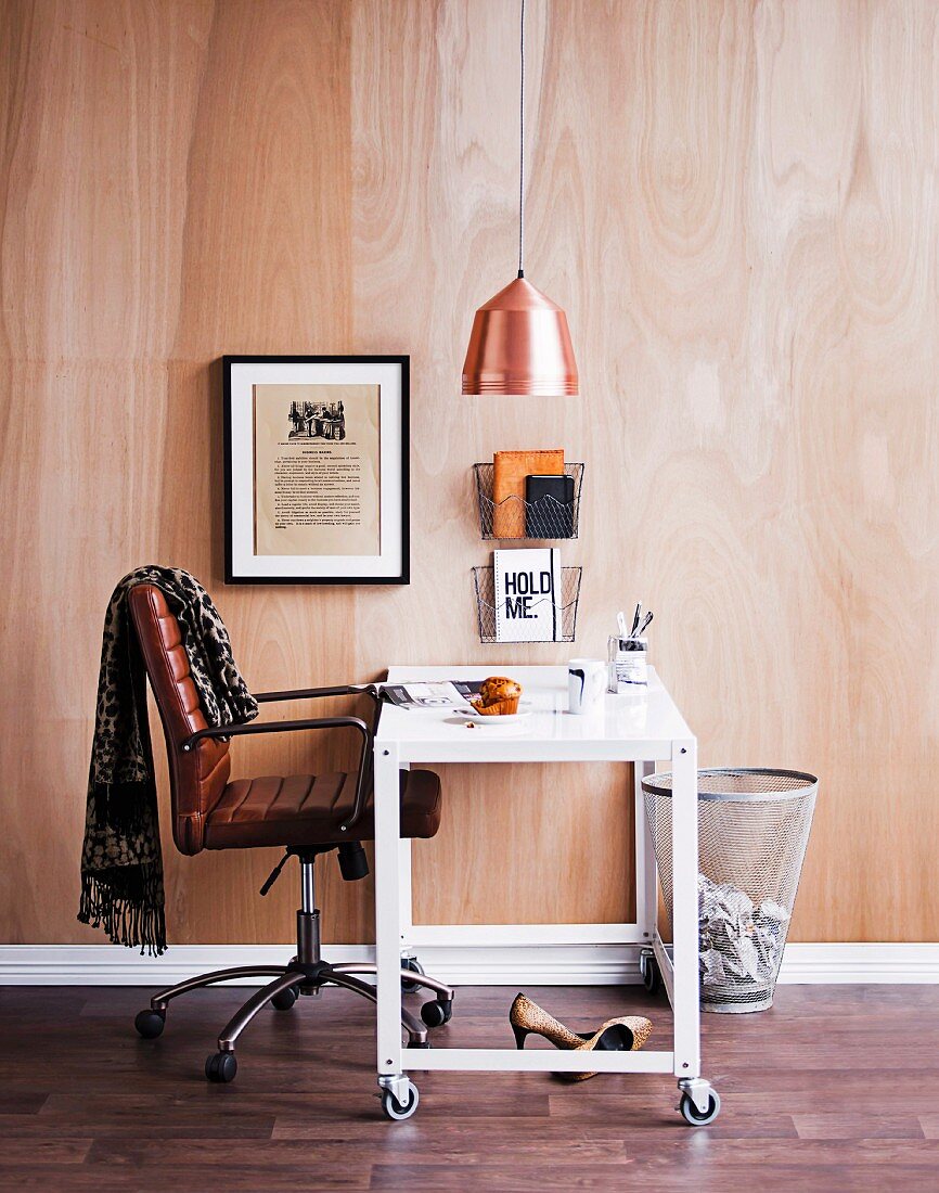 Bürostuhl und weisser Tisch mit Rollen unter Hängeleuchte mit Kupferschirm vor Holzwand