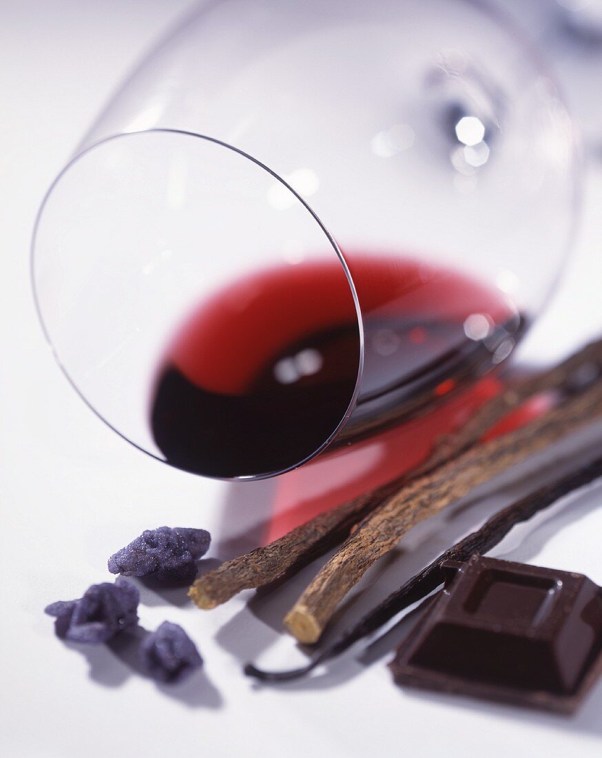 A glass of red wine lying on its side, with candied violets, dark chocolate, a vanilla pod and liquorice roots