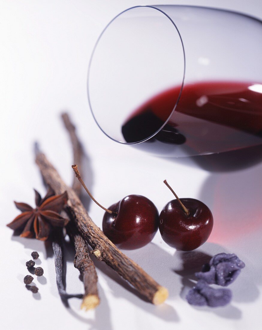 A glass of red wine lying on its side, with spices, cherries and candied violets