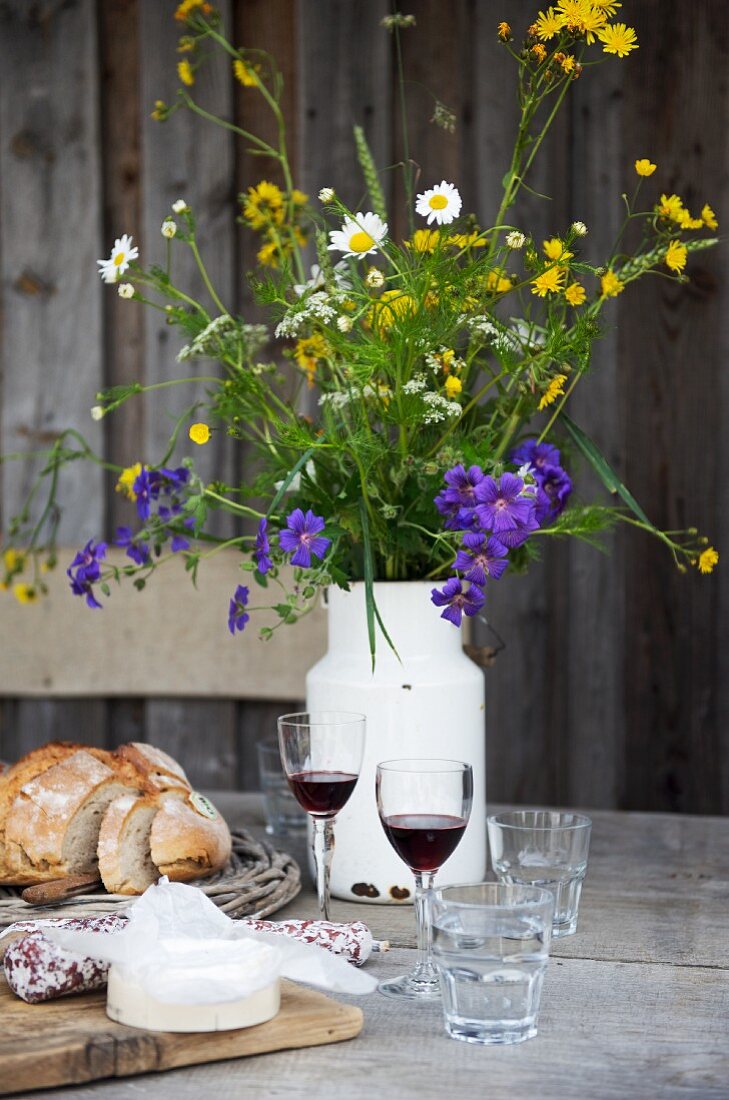 Cheese, bread, two glasses of red wine and a milk churn of wild flowers on a rustic wooden table