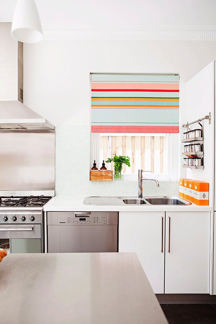 View over a table to a kitchen sink at a window, with a half-closed Roman blind with colorful stripes next to a gas oven in a modern kitchen