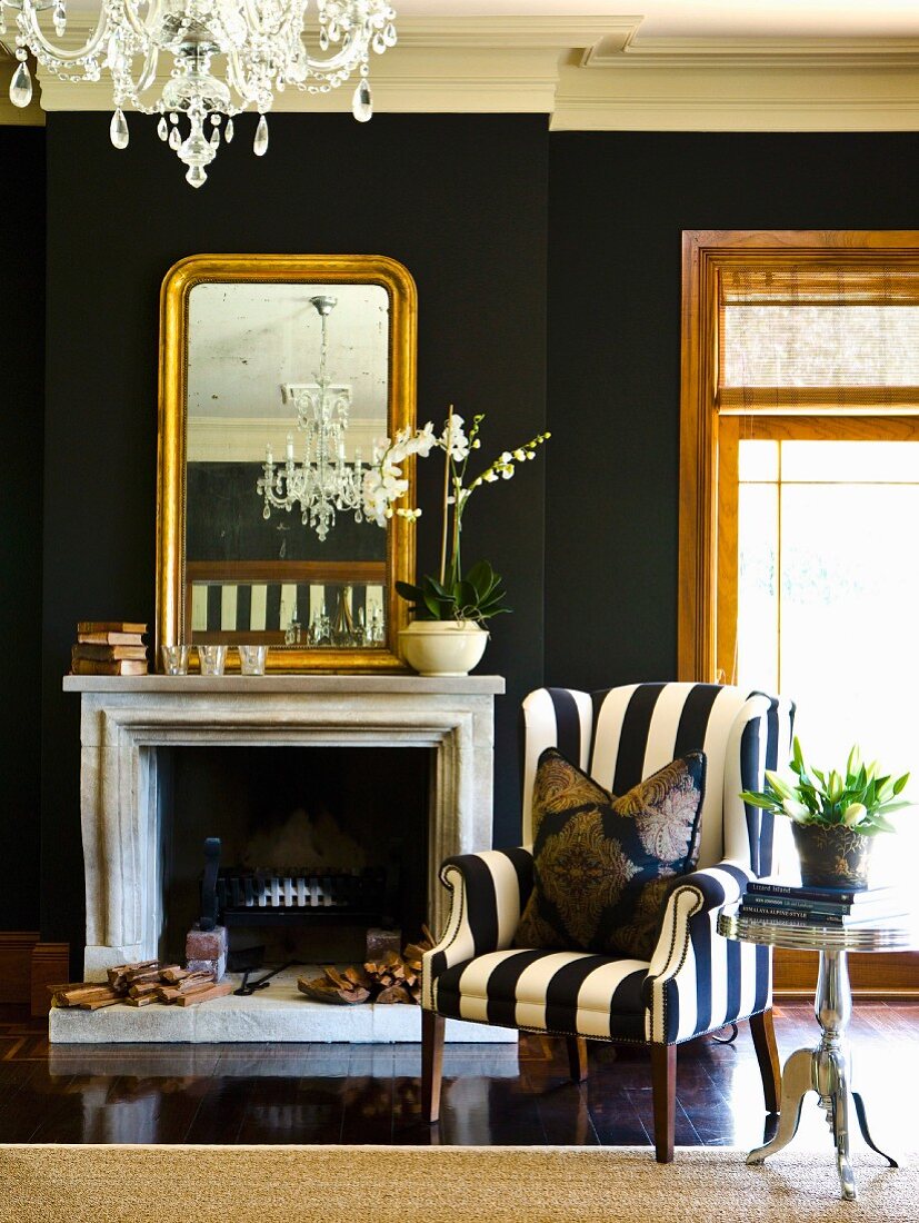 Black and white armchair next to an open fireplace and a gold framed mirror on a black wall