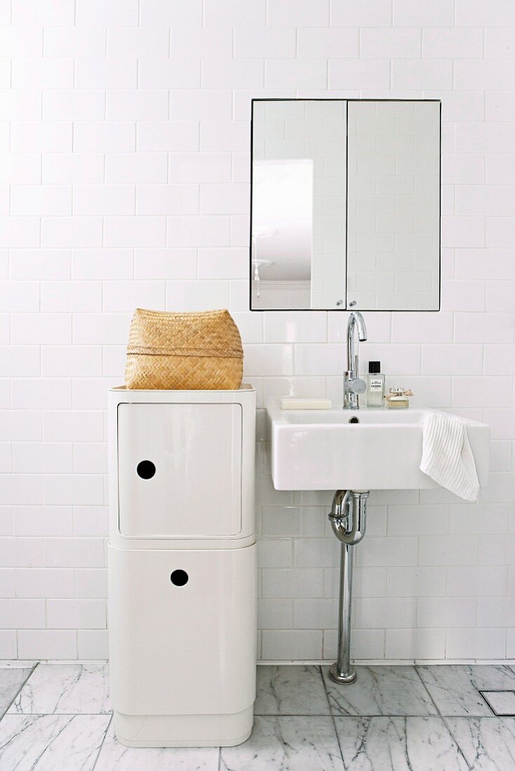 Half-height, white designer storage cabinet next to minimalist washstand against white tiled wall with integrated mirrored cabinet
