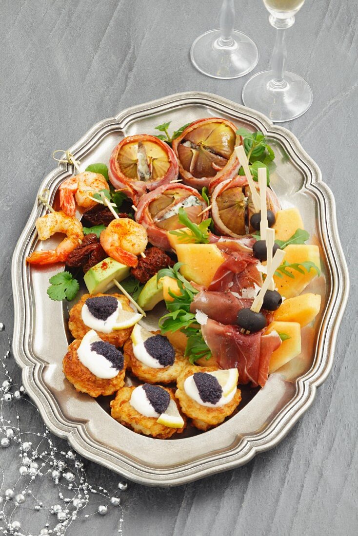 A platter of appetisers for Christmas, with melon, ham, potato fritters with caviar, figs with blue cheese, and prawn skewers