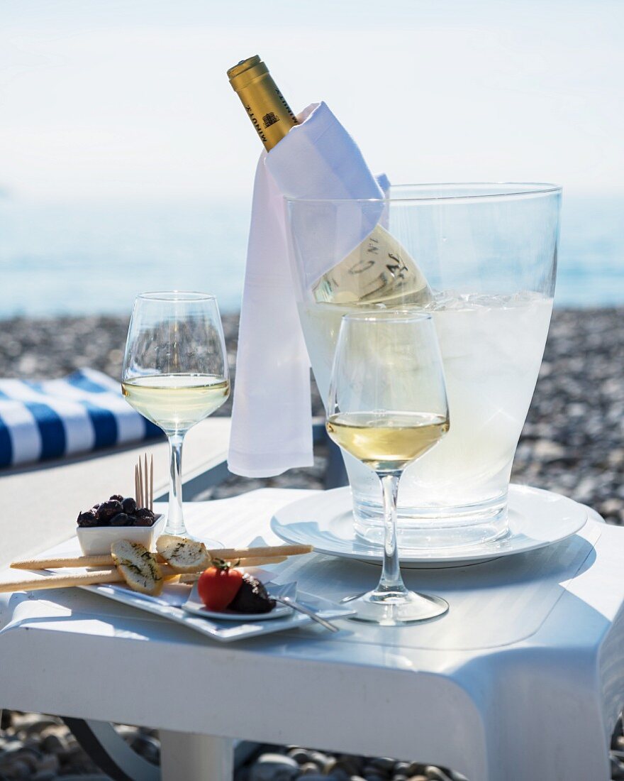 A table laid for two at the beach, with appetisers, glasses of white wine and a bottle of wine in a chiller