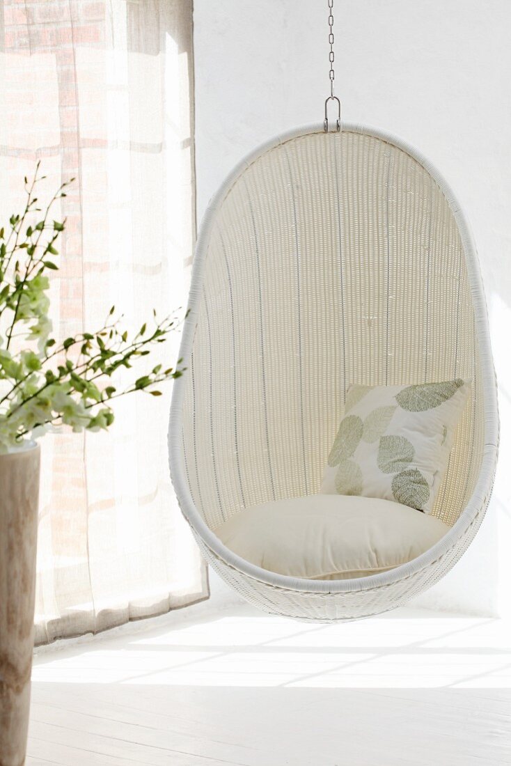 Comfortable, white wicker hanging chair in corner of room next to window with closed, translucent curtains and pattern of light and shadow on floor