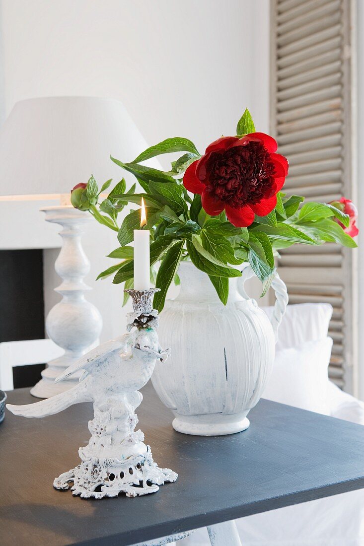Vase mit roter Blume und Porzellankerzenständer vor Tischlampe auf dunkler Ablage